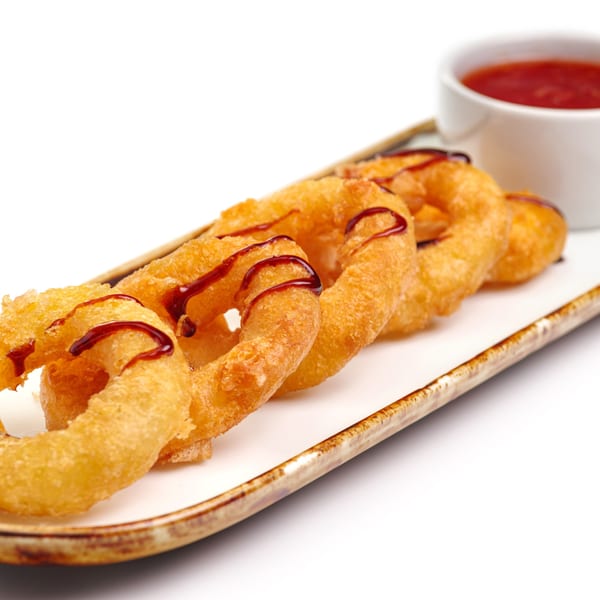 Golden-brown onion rings drizzled with sauce, served on a white rectangular plate with a side of red dipping sauce.