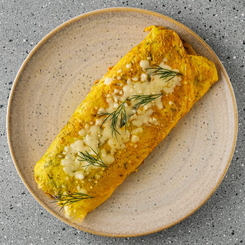 Golden omelet topped with melted cheese and fresh dill sprigs on a speckled plate.