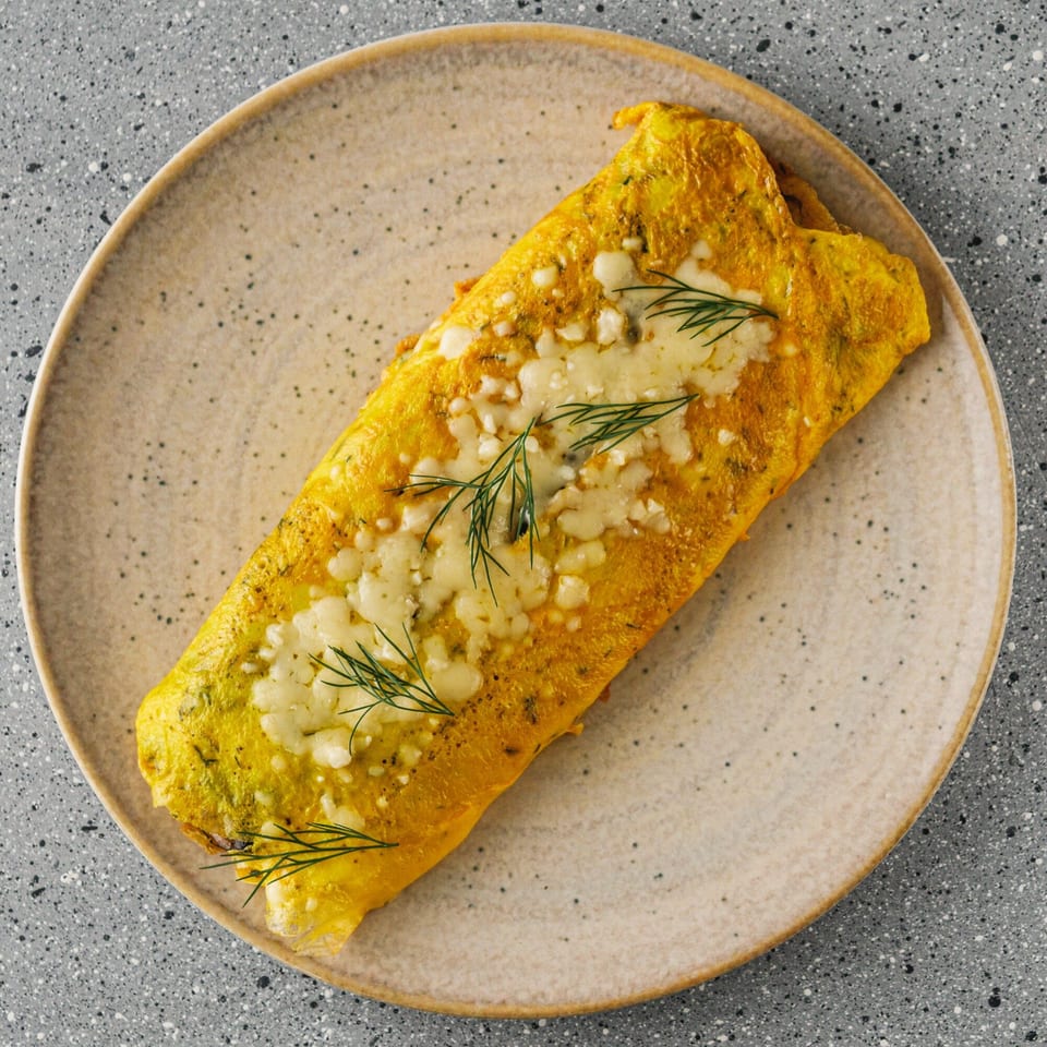 A golden omelet topped with melted cheese and fresh dill sprigs on a speckled plate.
